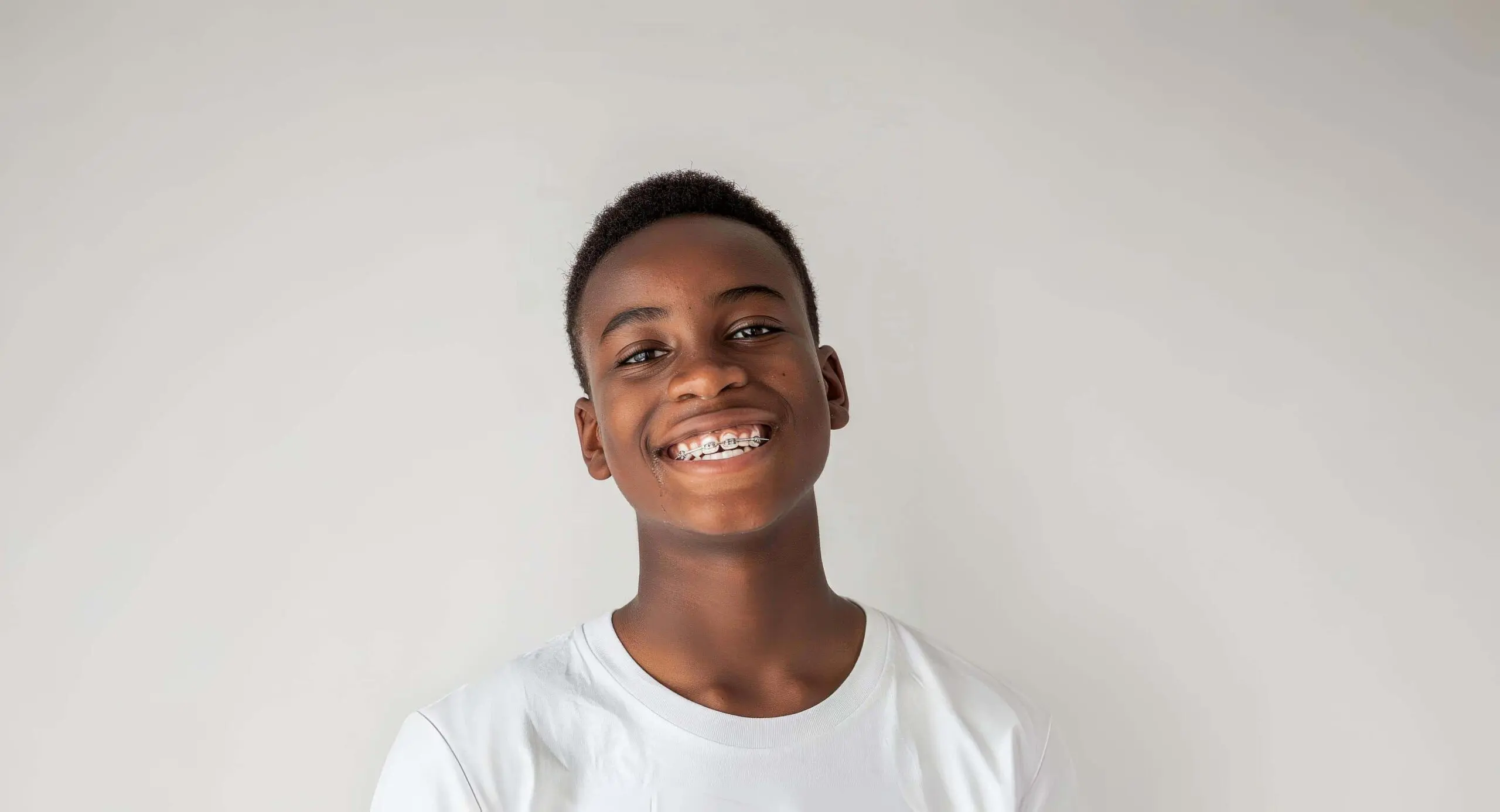 A smiling teen with traditional braces and a white t-shirt stands against a light background at BeSpoke Orthodontics in Burtonsville, MD.