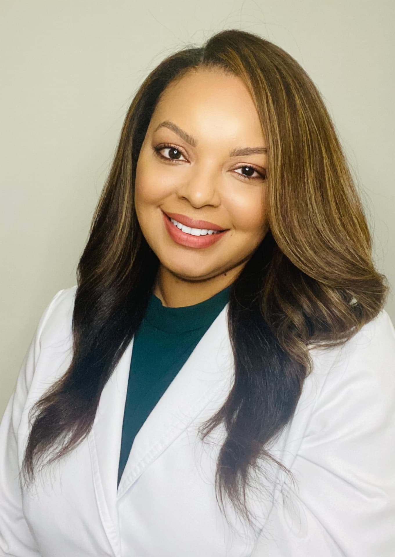 At Bespoke Orthodontics in Burtonsville, MD, Dr. Latoya Callahan—wearing a white lab coat and dark green top, with long brown hair—smiles at the camera against a plain background.