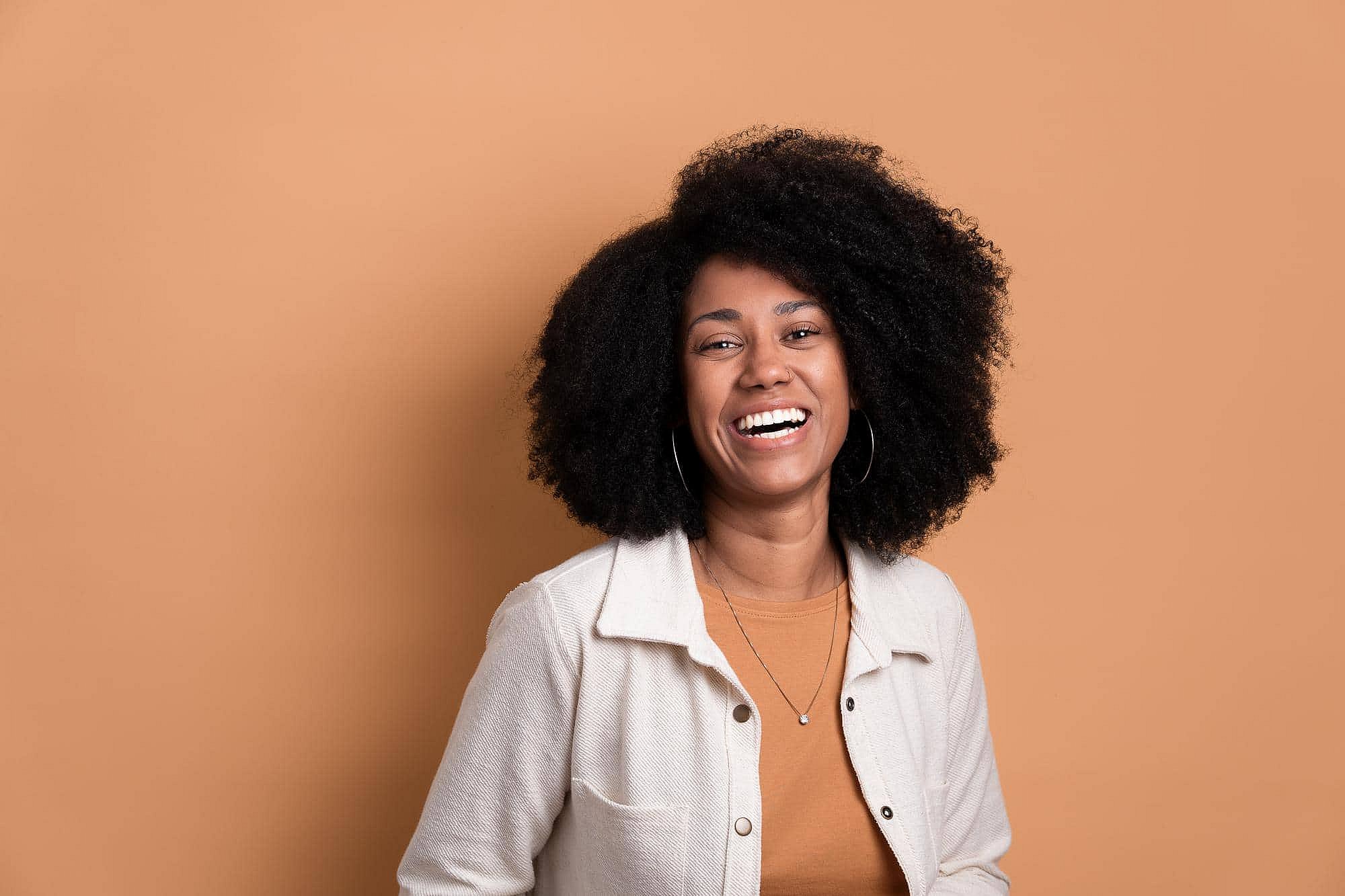 Smiling woman with curly hair and metal braces at BeSpoke Orthodontics in Burtonsville, MD, stands against a beige background.