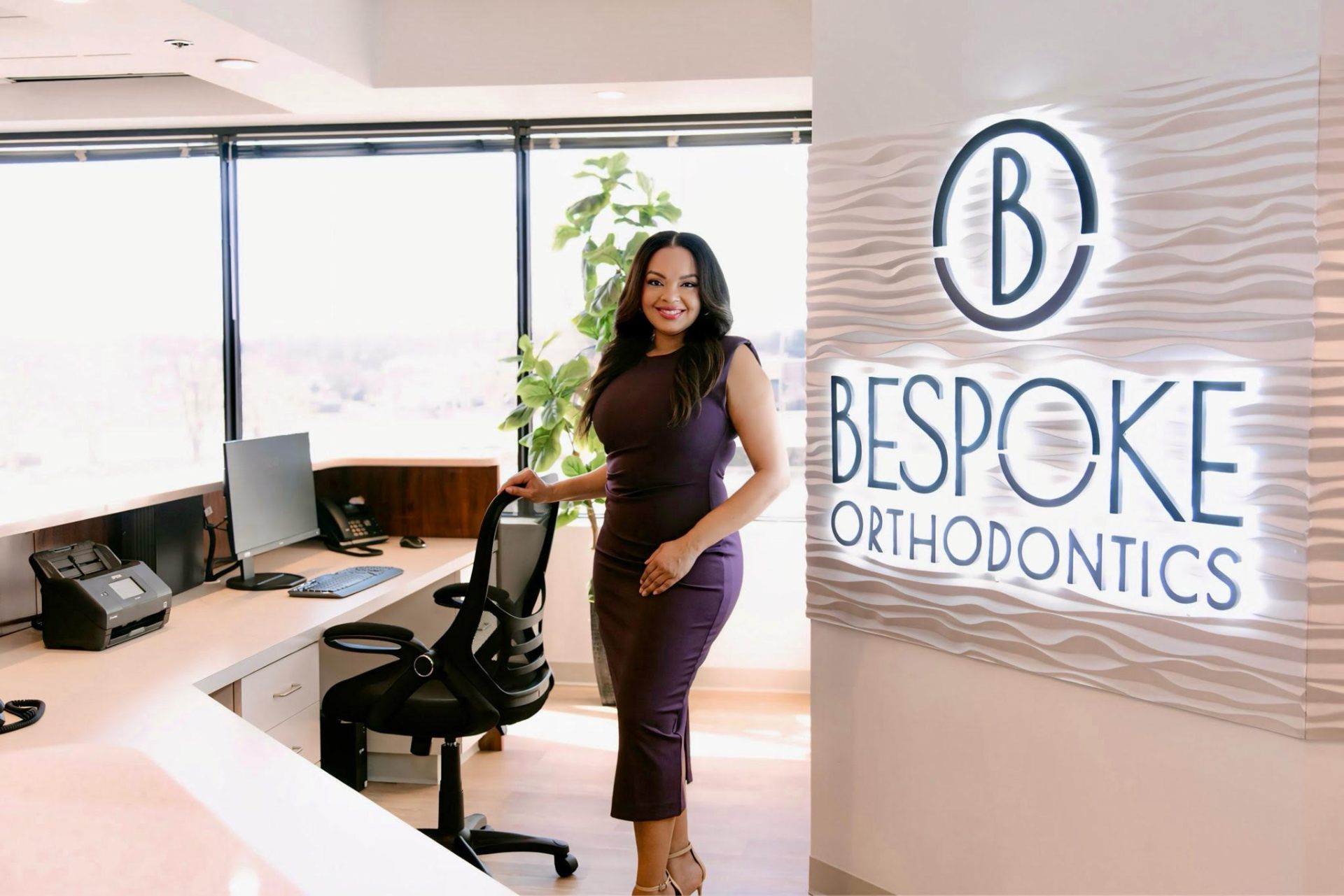 In a modern office in Burtonsville, MD, a woman in a purple dress stands by an office chair near the BeSpoke Orthodontics sign.