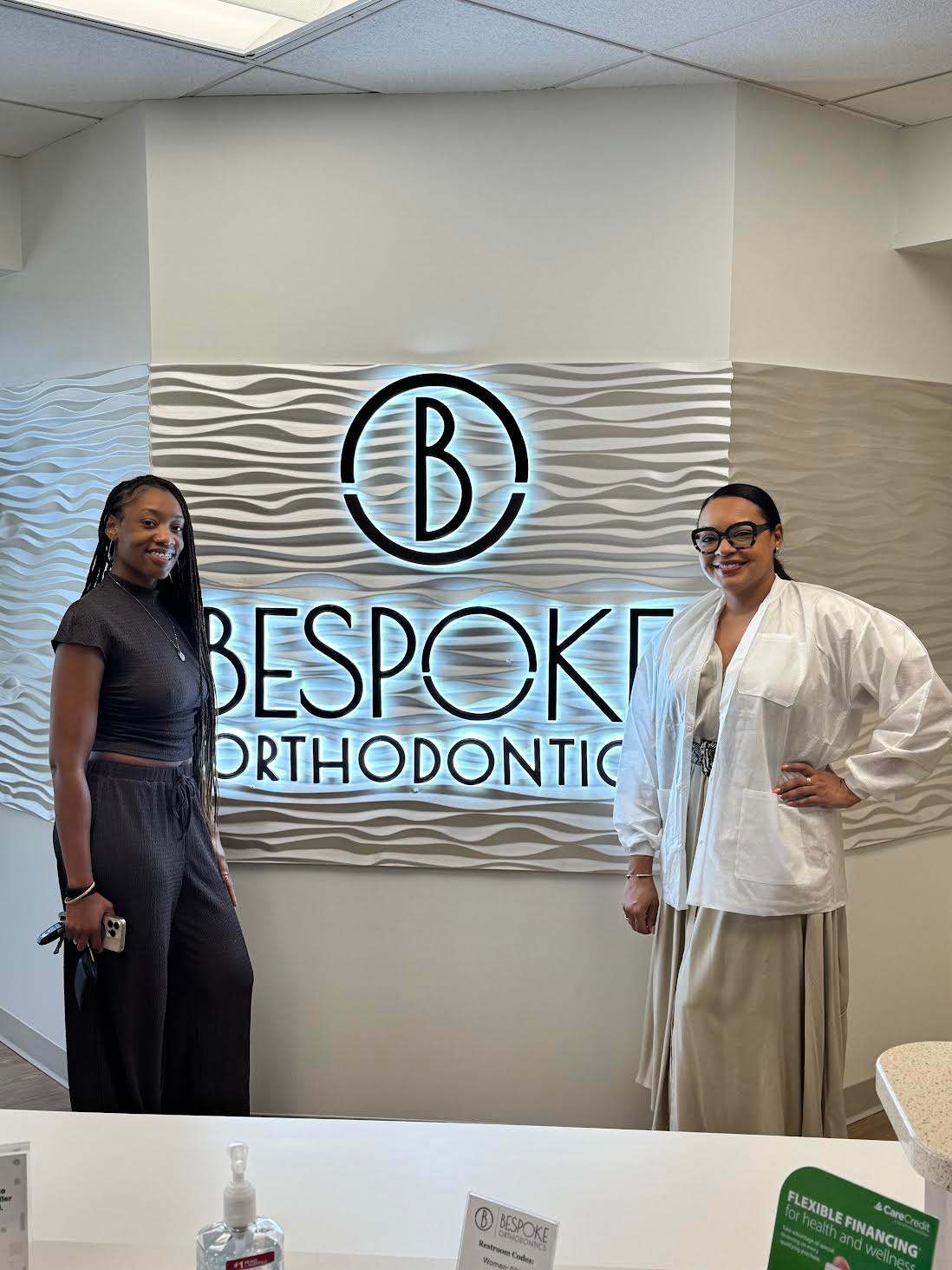 Two women stand in a modern office, with a wall sign for BeSpoke Orthodontics in Burtonsville, MD visible behind them.