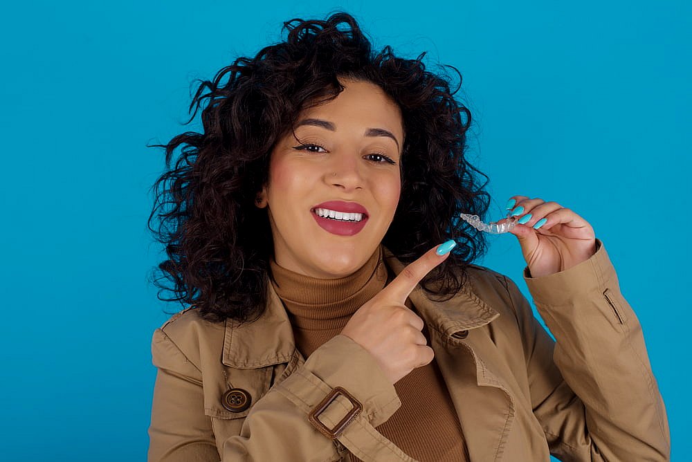 Smiling woman with curly hair in a brown coat points at an Invisalign aligner; BeSpoke Orthodontics in Burtonsville, MD.