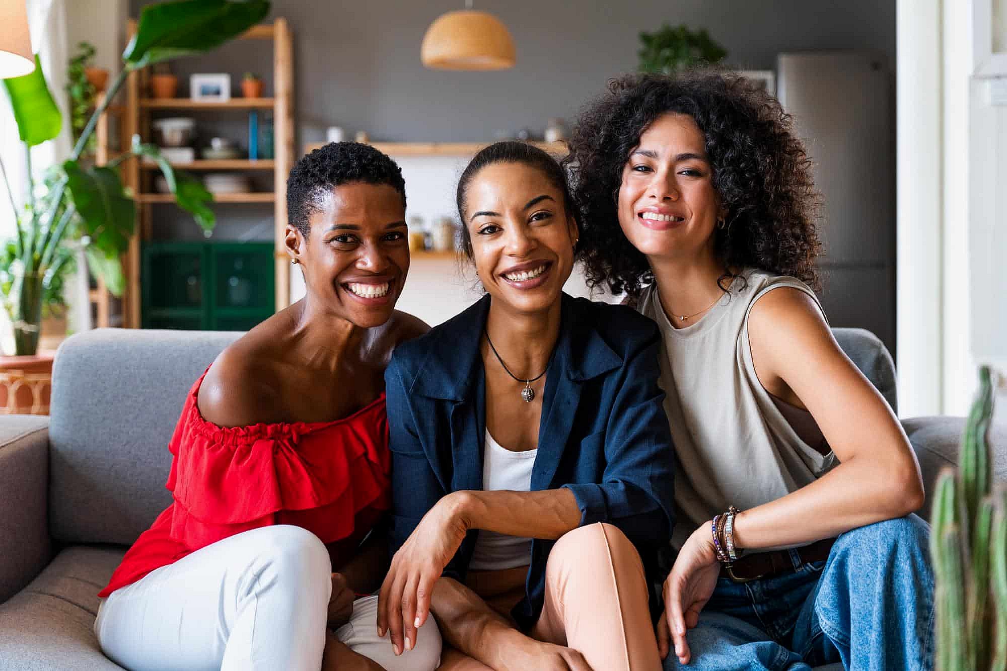 Three women with confident, bright smiles from BeSpoke Orthodontics in Burtonsville, MD sit together on a modern living room couch.