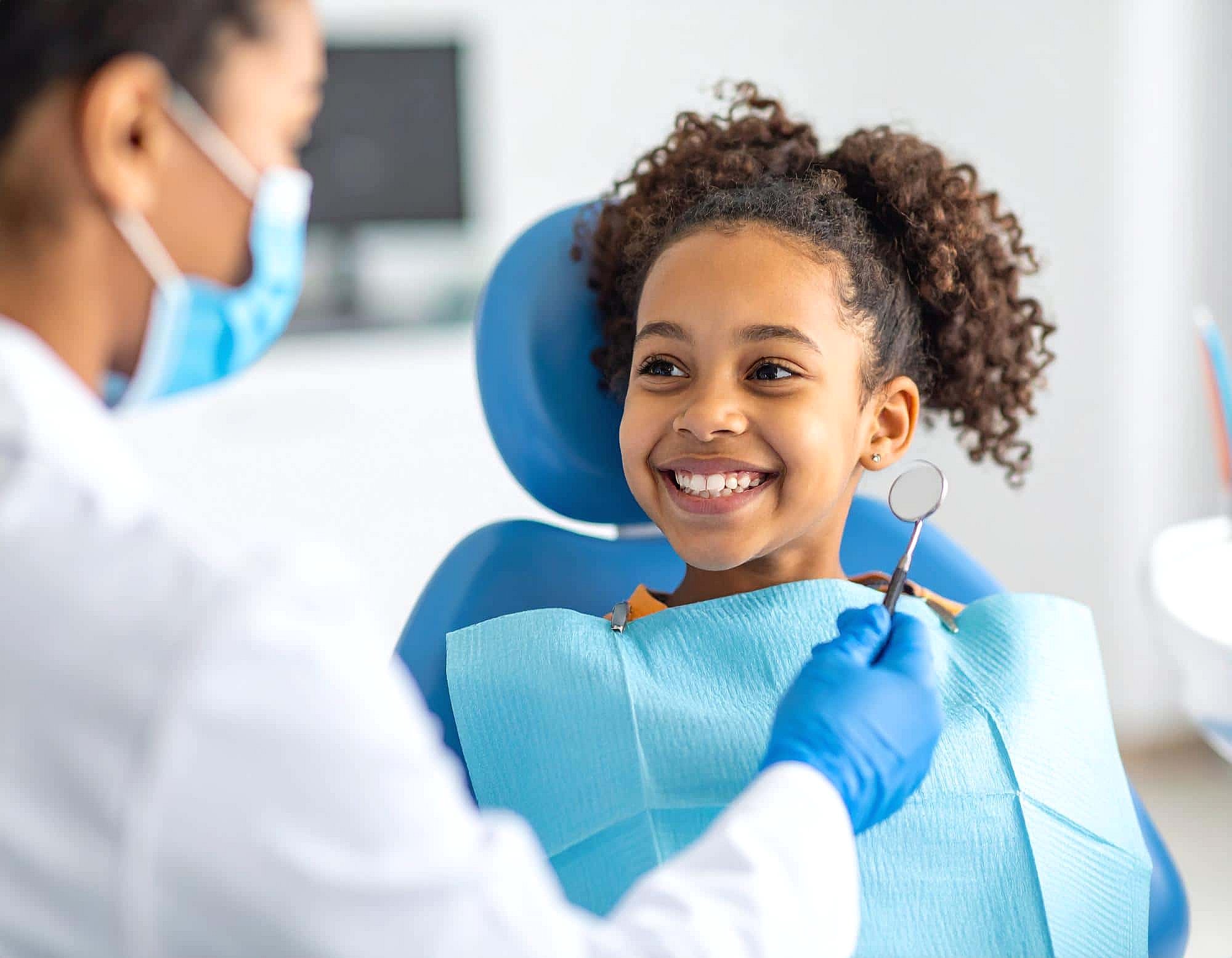 A young girl smiles in the dental chair at BeSpoke Orthodontics in Burtonsville, MD as the orthodontist holds a dental mirror.