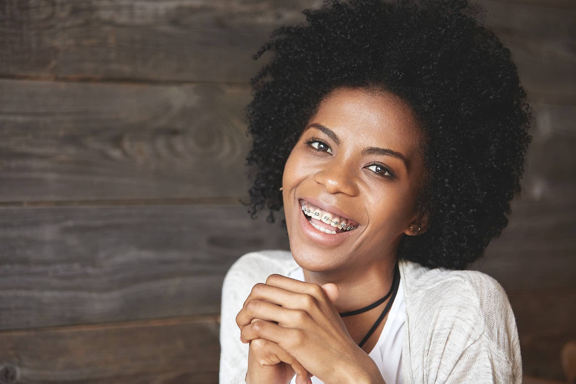 Smiling young woman with curly hair and metal braces, at BeSpoke Orthodontics in Burtonsville, MD, sits by a wooden wall.