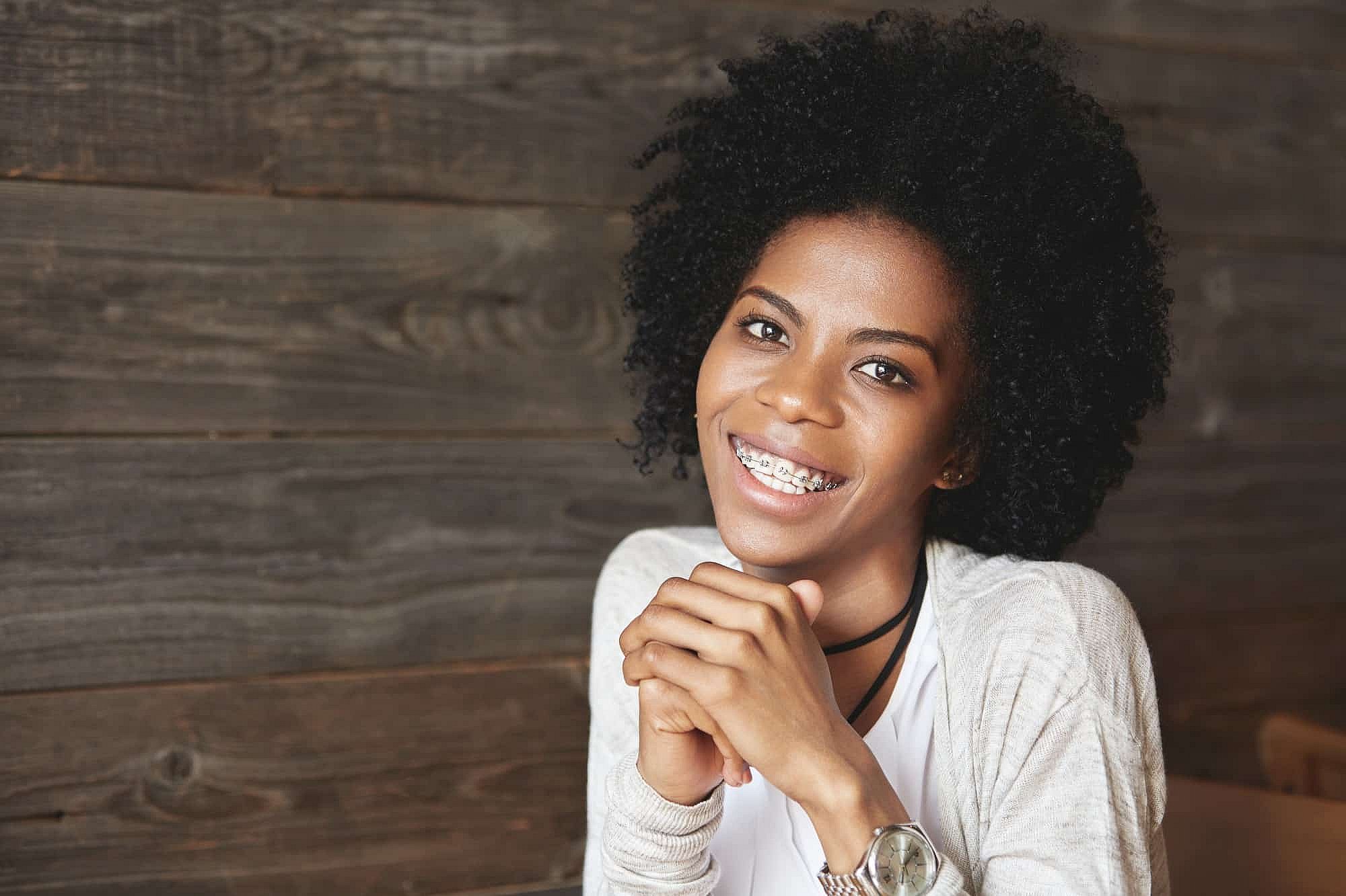 Smiling with metal braces, a young woman sits at a wooden table at BeSpoke Orthodontics in Burtonsville, MD.