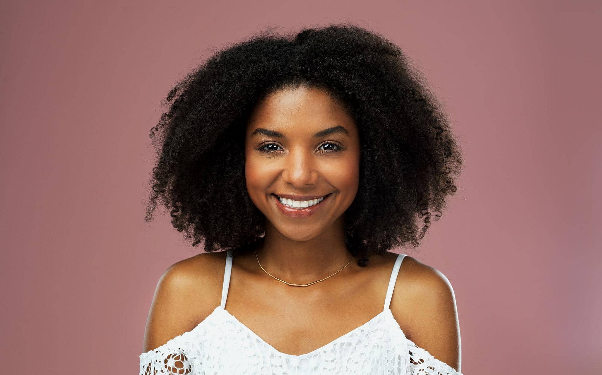 Smiling woman with curly hair and metal braces represents BeSpoke Orthodontics in Burtonsville, MD, on a pink background.