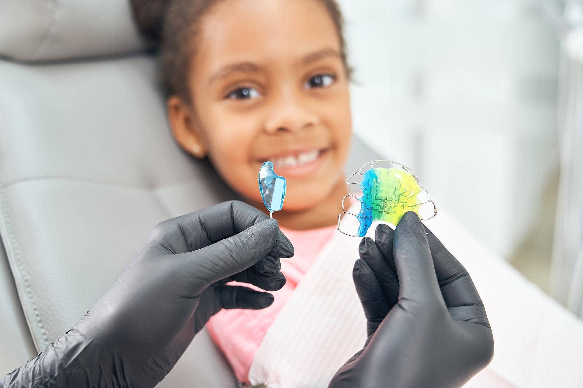 At BeSpoke Orthodontics in Burtonsville, MD, a child smiles in the dental chair as colorful retainers are displayed by a gloved professional.