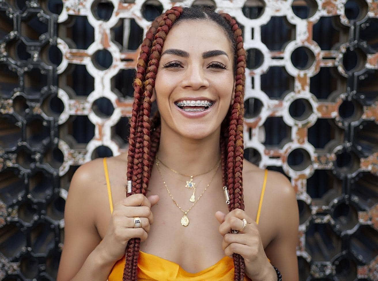 Smiling at the camera, a woman with red braids and braces from BeSpoke Orthodontics in Burtonsville, MD stands against a geometric background.