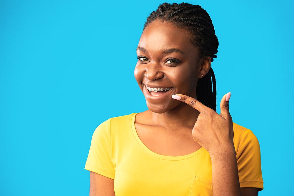 Smiling in a yellow shirt, a young woman points to her braces at BeSpoke Orthodontics in Burtonsville, MD, on a blue background.
