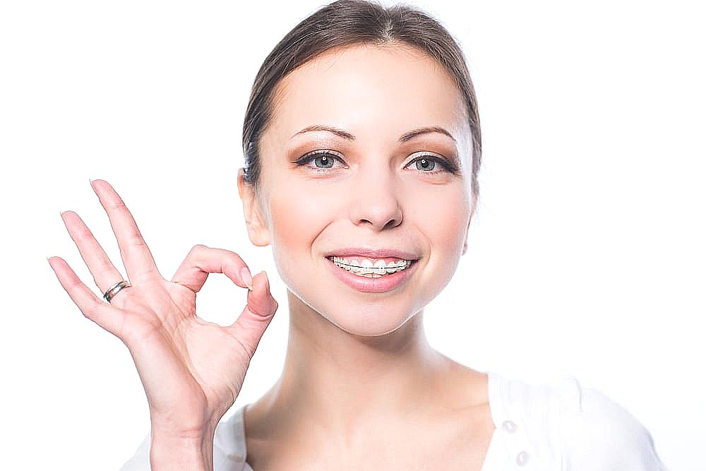 Smiling woman with clear braces gives an "OK" gesture, representing BeSpoke Orthodontics in Burtonsville, MD, on a white background.