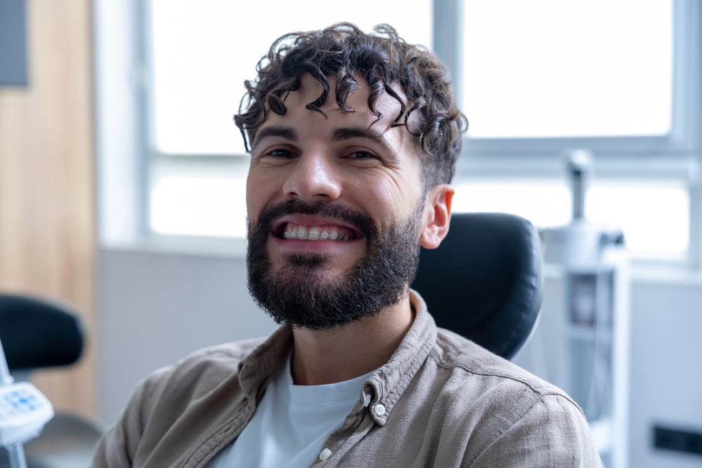A bearded man with curly hair and clear braces smiles in a dental chair at BeSpoke Orthodontics in Burtonsville, MD.