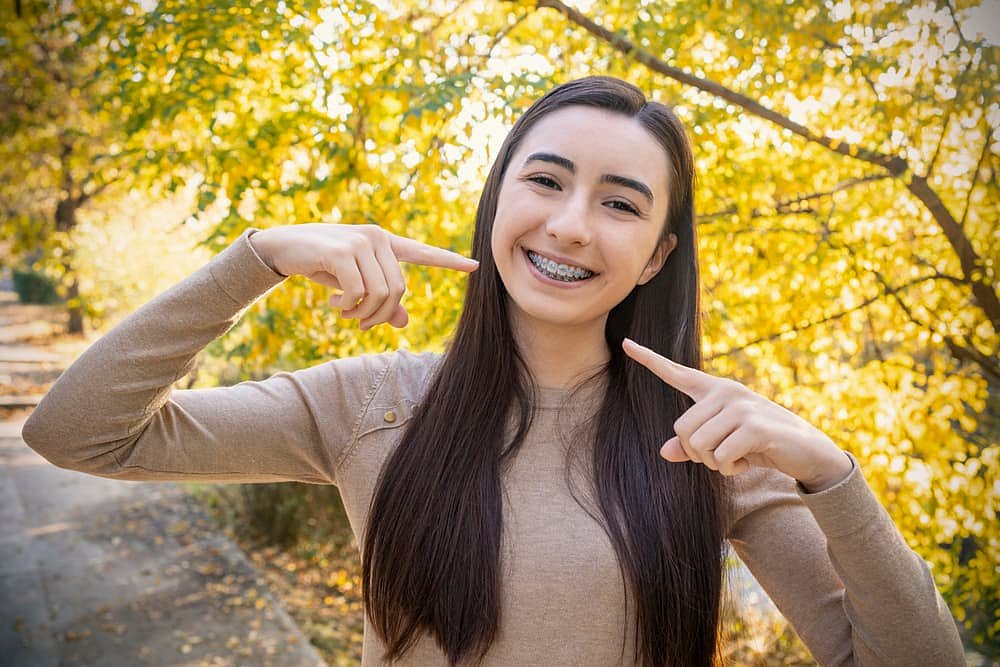 Smiling young woman with metal braces points to her teeth outdoors, promoting BeSpoke Orthodontics in Burtonsville, MD, with fall leaves.