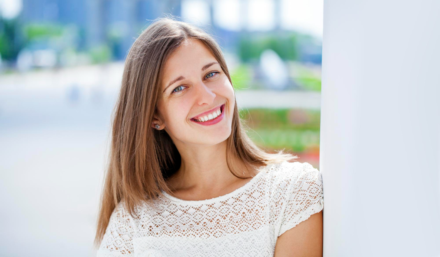 Smiling young woman with straight brown hair and a white lace top outdoors by a white wall represent orthodontic treatment options for Adult Orthodontics at BeSpoke Orthodontics in Burtonsville, MD.