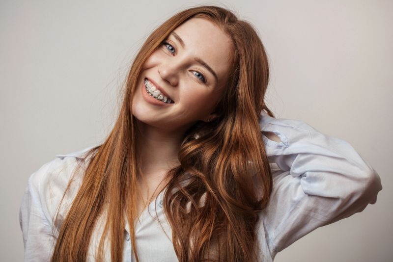 Smiling young woman with red hair and clear braces, wearing a blue shirt; BeSpoke Orthodontics in Burtonsville, MD.