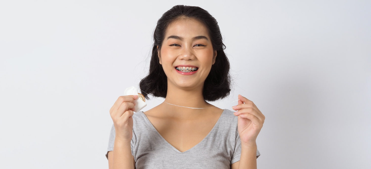 Smiling young woman with braces holds dental floss, promoting BeSpoke Orthodontics in Burtonsville, MD, highlighting Braces Cost; plain white background.