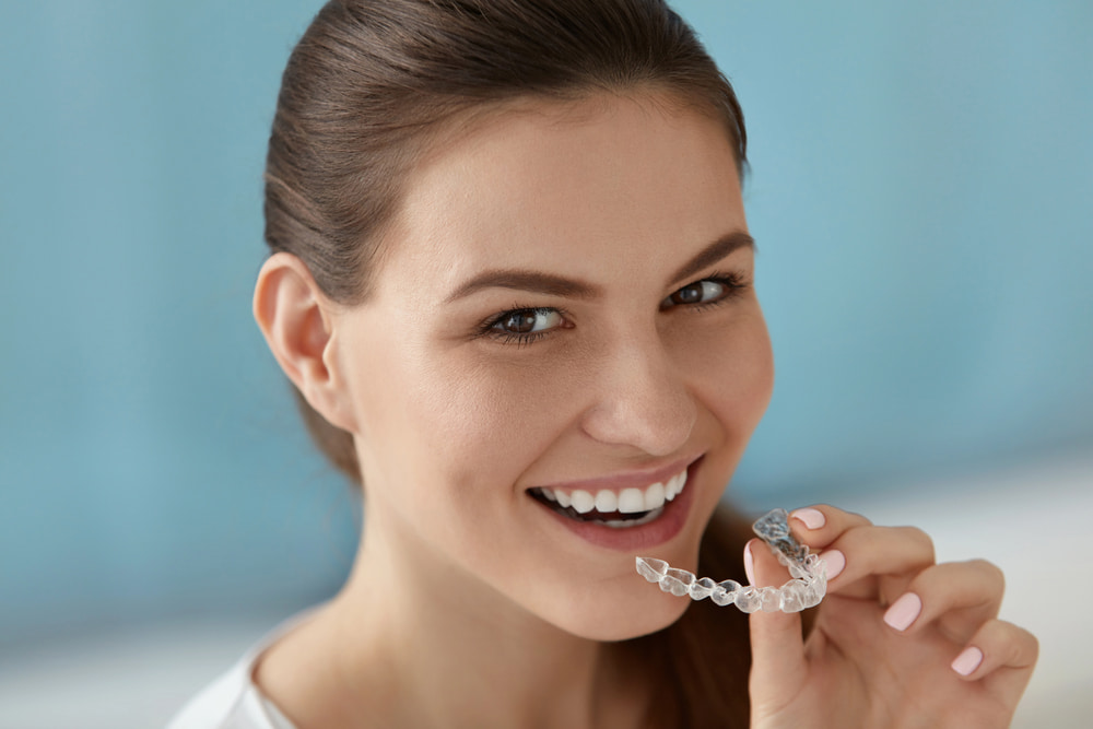A woman smiles and holds a clear aligner near her mouth, highlighting BeSpoke Orthodontics' Invisalign Payment Plan in Burtonsville, MD.
