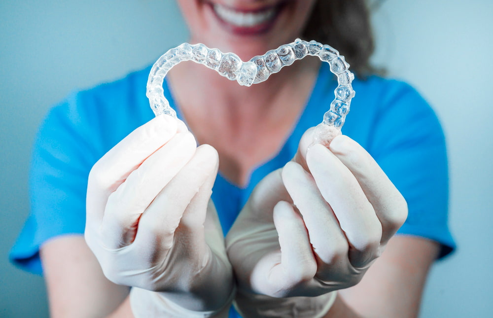 Gloved hands holding two clear aligners in a heart shape at BeSpoke Orthodontics in Burtonsville, MD, with a smiling face blurred in the background, highlighting Invisalign cost.