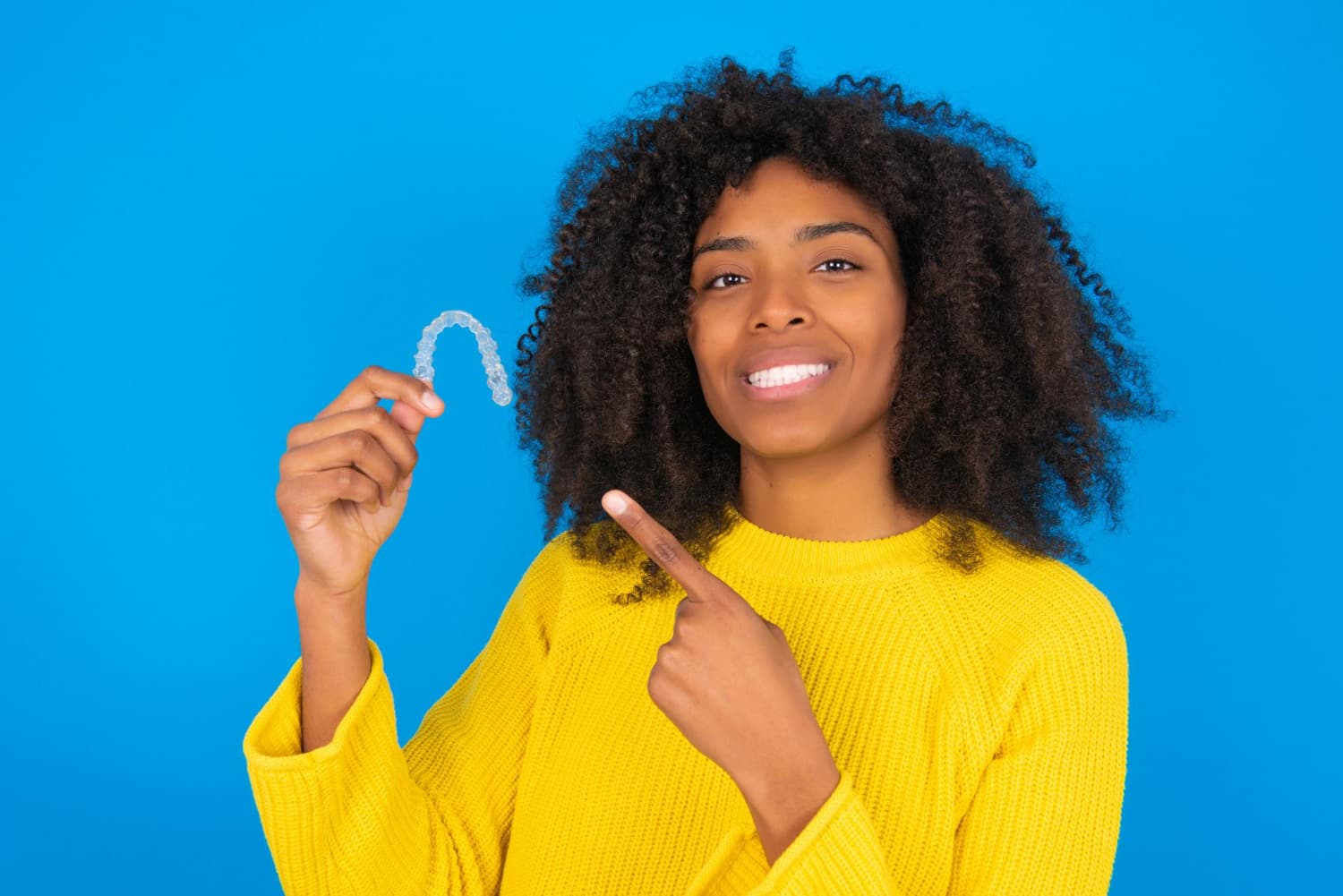 Smiling woman with curly hair holds Invisalign Clear Aligners at BeSpoke Orthodontics in Burtonsville, MD by a glass wall.